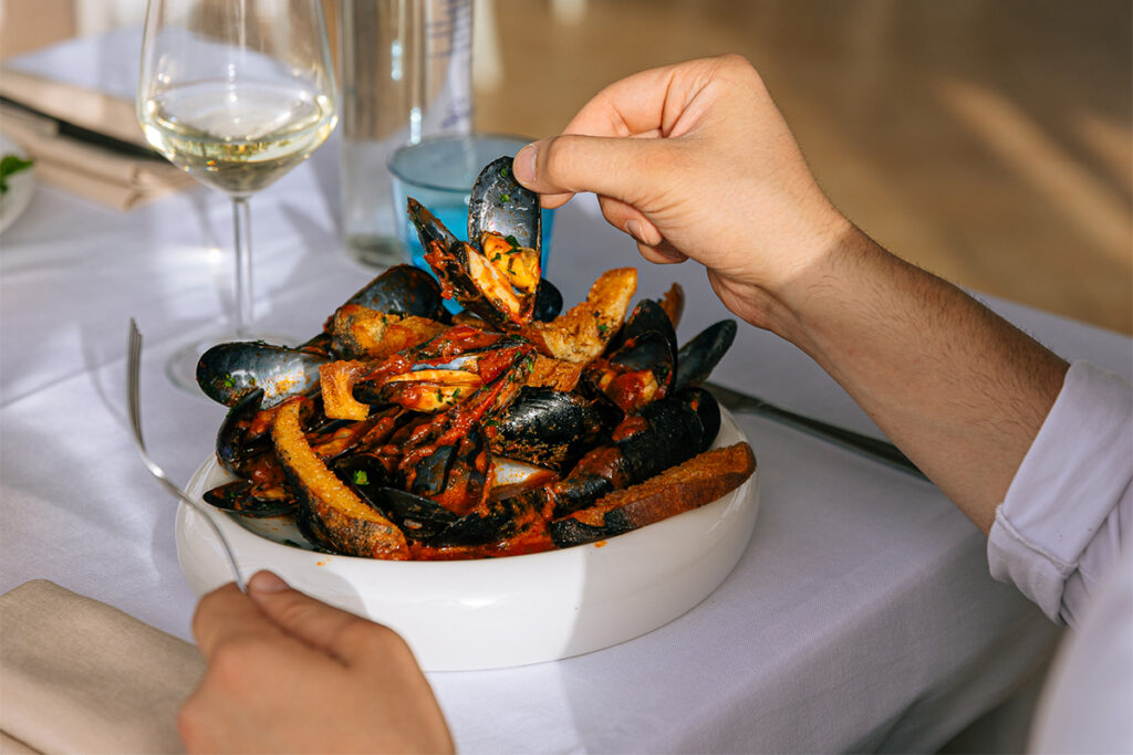 Sautè di cozze con crostini, servito in una ciotola bianca mentre due persone si servono