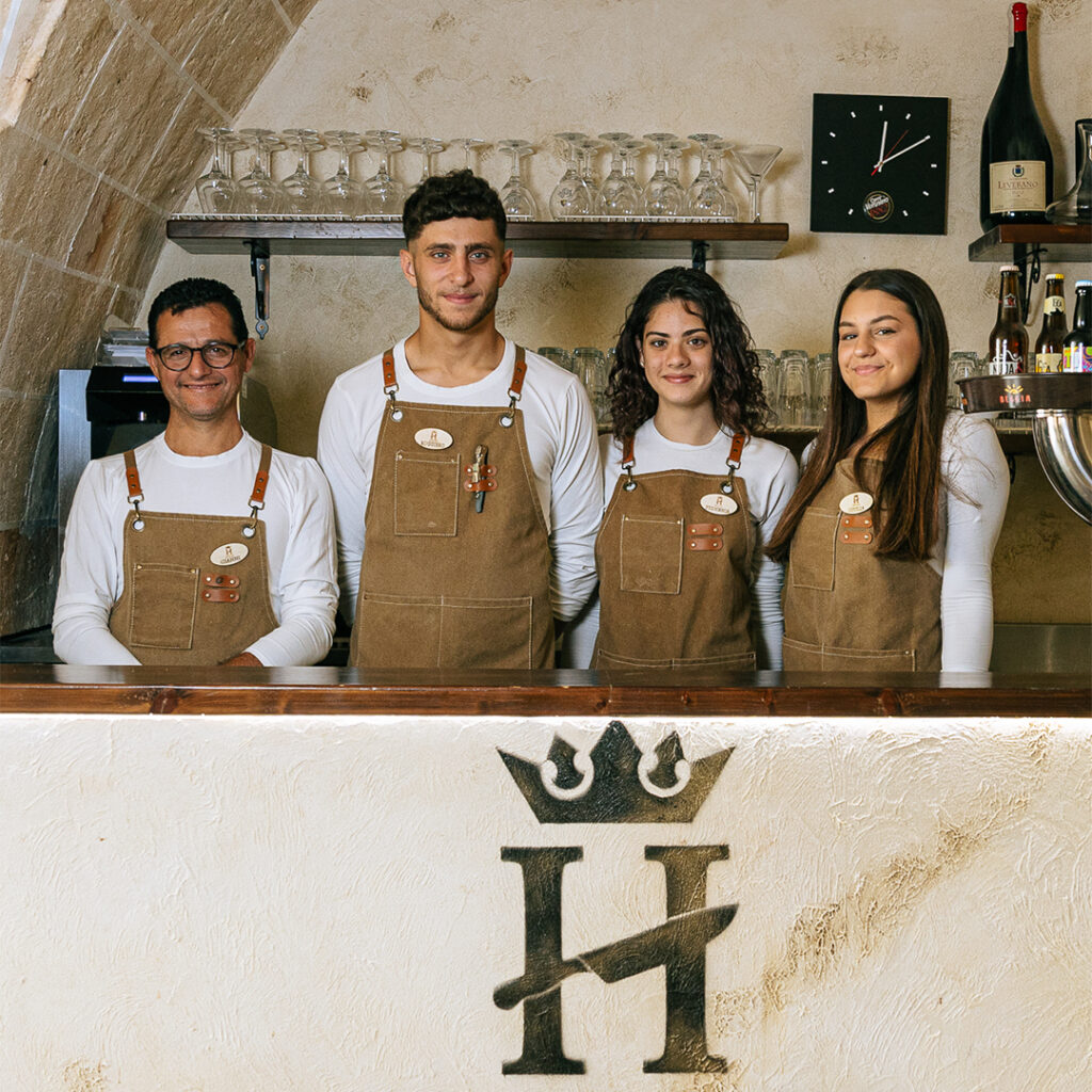La squadra di House dietro il bancone, con i grembiuli color cuoio e il monogramma H con la corona inciso nel muro