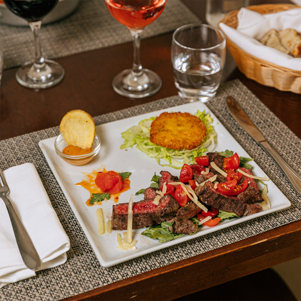 Carne alla griglia servita con rucola, pomodorini e scaglie di grana su piatto quadrato bianco
